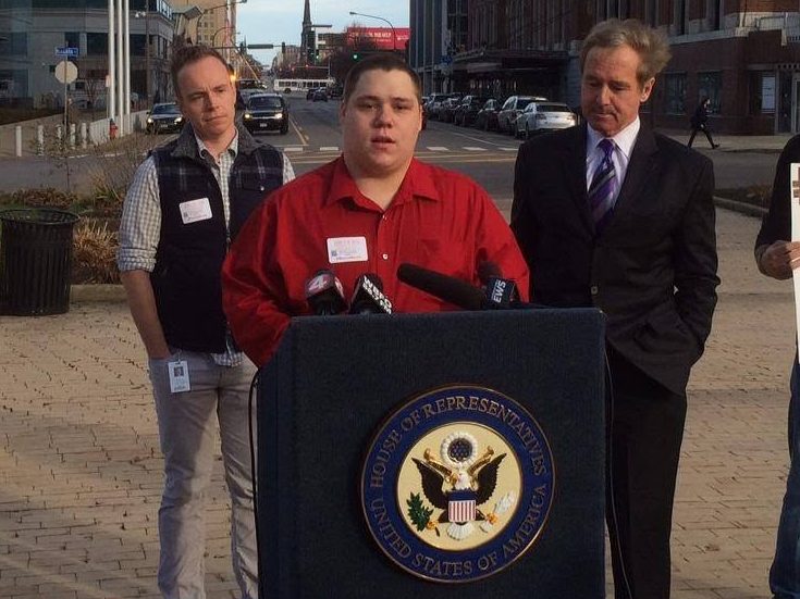 A person in a red shirt speaks at a podium with a seal, flanked by two men.